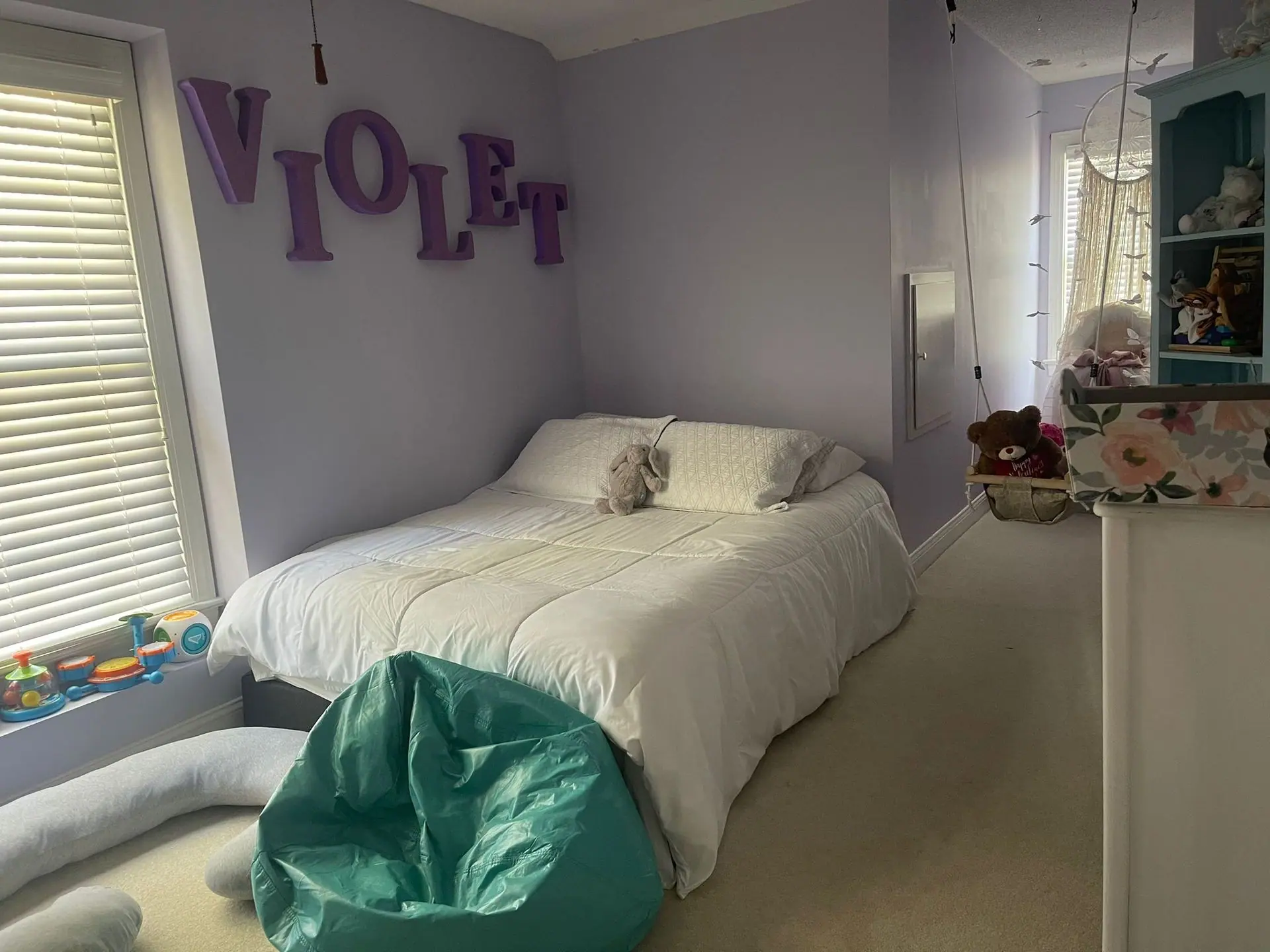 A bedroom with light purple walls, a bed with white bedding, a teal beanbag chair, and the name