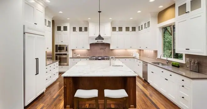A spacious, modern kitchen with white cabinets, a large marble-topped island, wooden floors, and sta…